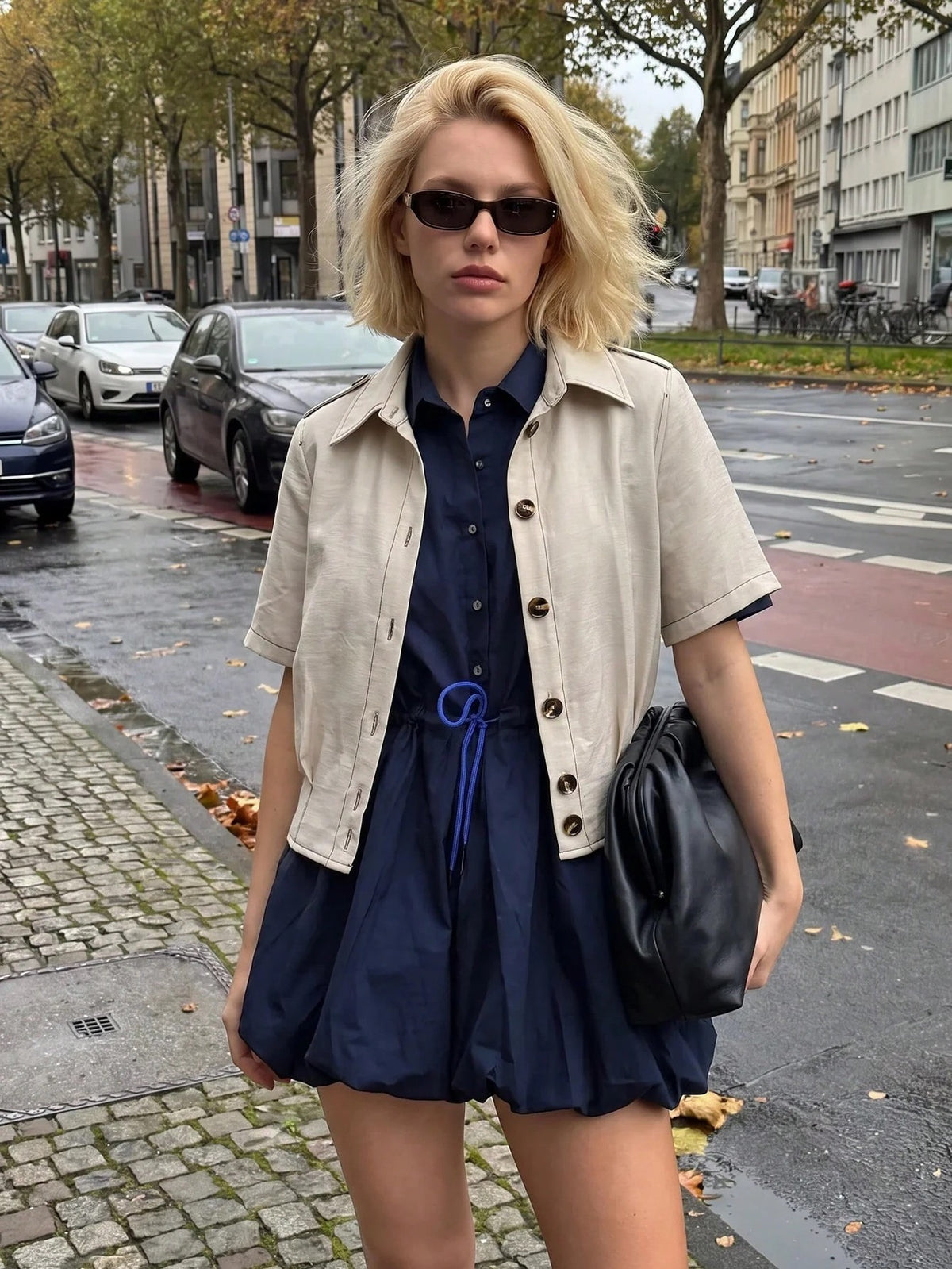 A blonde woman wearing a khaki short-sleeve lapel crop jacket over a dark blue dress while walking outdoors on a city street