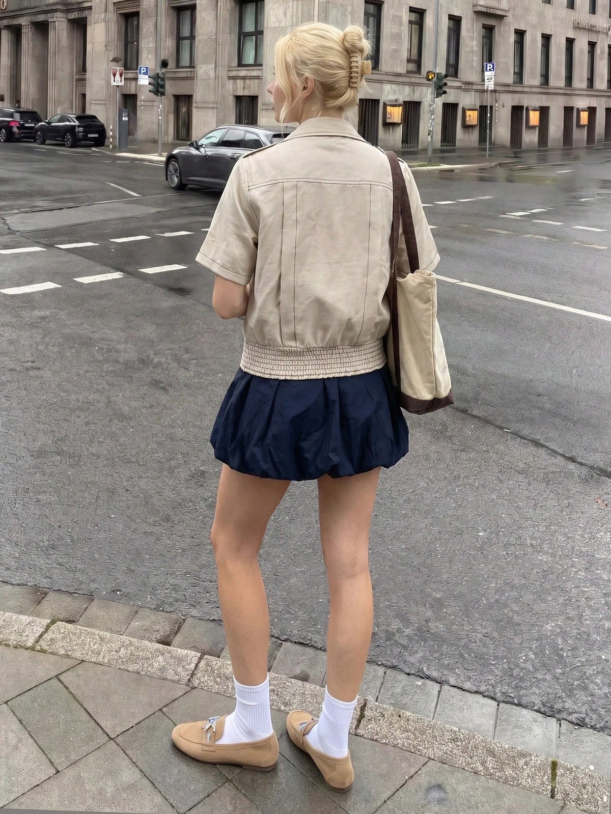 A blonde woman wearing a khaki short-sleeve lapel crop jacket over a dark blue dress while walking outdoors on a city street