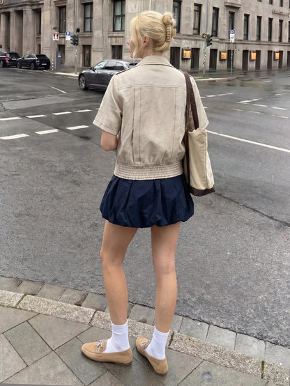 A blonde woman wearing a khaki short-sleeve lapel crop jacket over a dark blue dress while walking outdoors on a city street