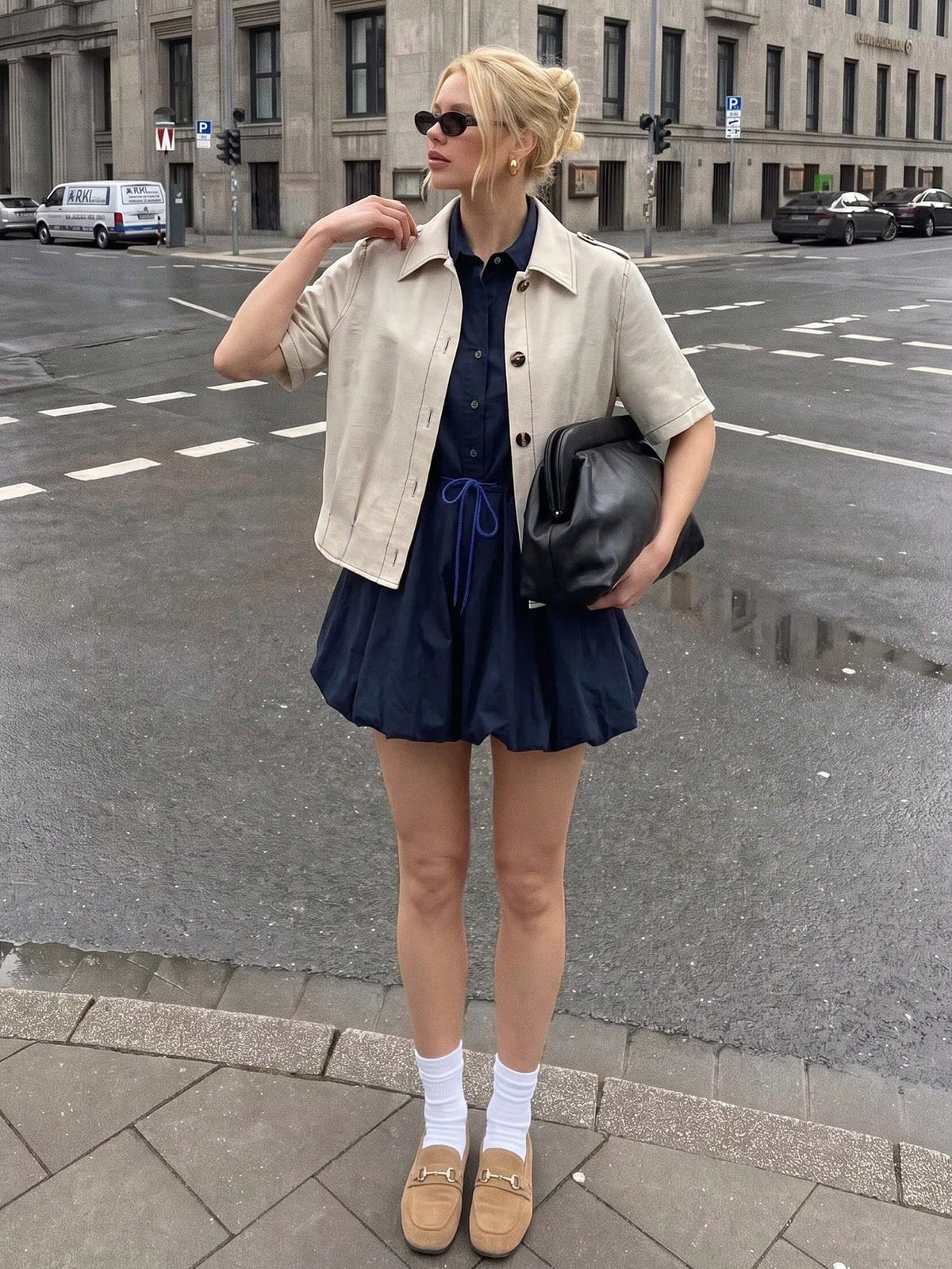 A blonde woman wearing a khaki short-sleeve lapel crop jacket over a dark blue dress while walking outdoors on a city street