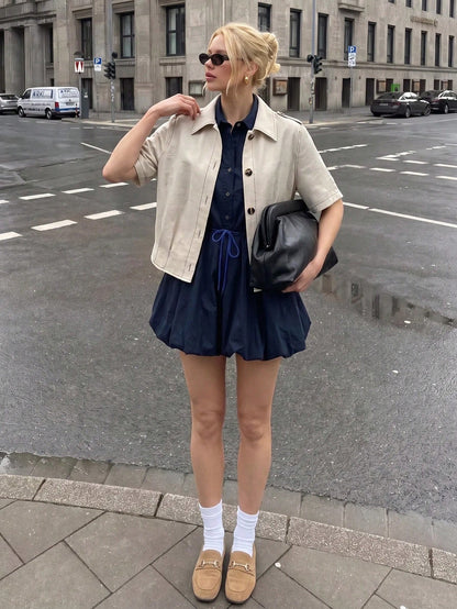 A blonde woman wearing a khaki short-sleeve lapel crop jacket over a dark blue dress while walking outdoors on a city street