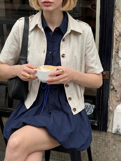 A blonde woman wearing a khaki short-sleeve lapel crop jacket over a dark blue dress while walking outdoors on a city street