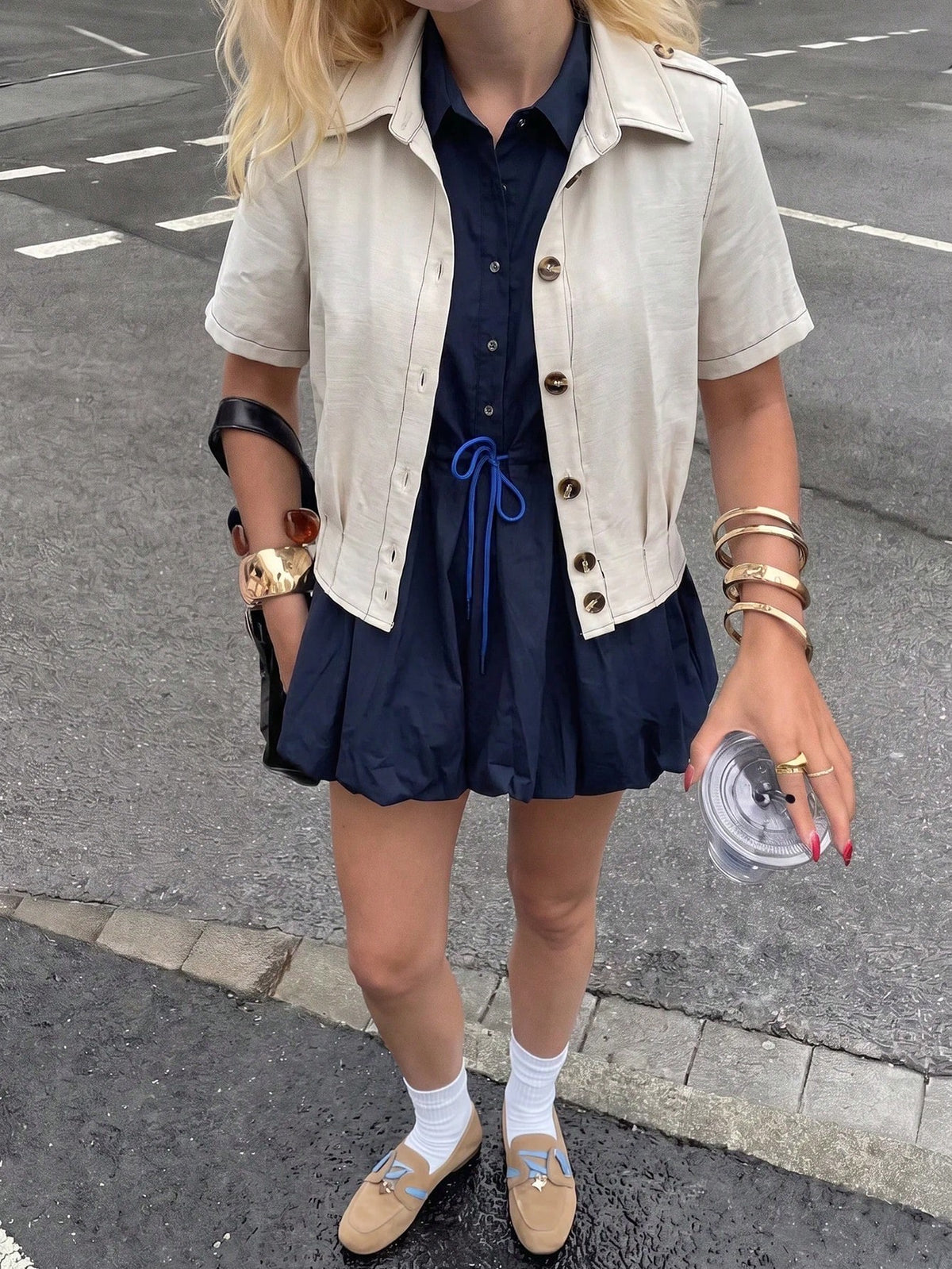 A blonde woman wearing a khaki short-sleeve lapel crop jacket over a dark blue dress while walking outdoors on a city street