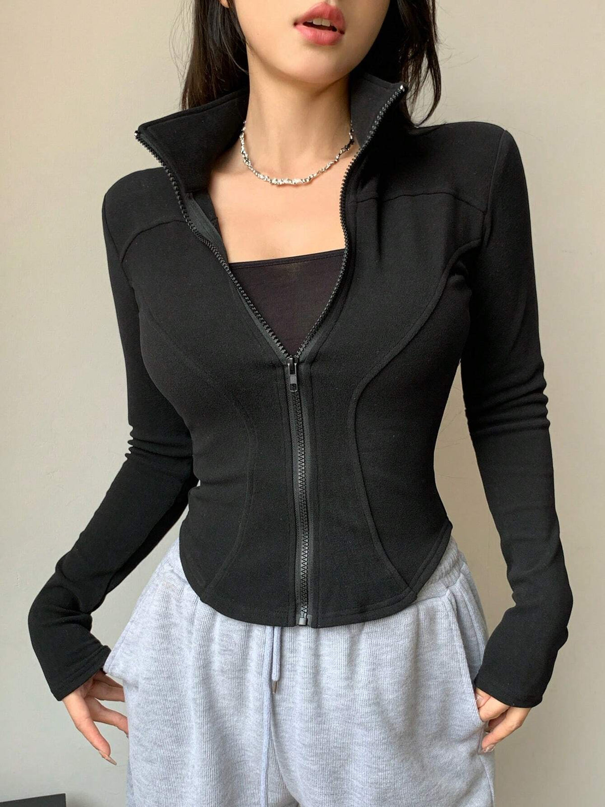 A woman wearing a black ribbed trim zip-up sweatshirt jacket with a high mock neck paired with light grey sweatpants