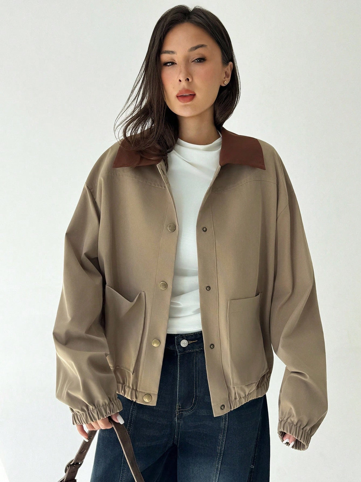 A woman wearing a khaki casual jacket with a contrasting brown lapel collar, drop shoulders, and front pockets paired with a white tee and dark jeans