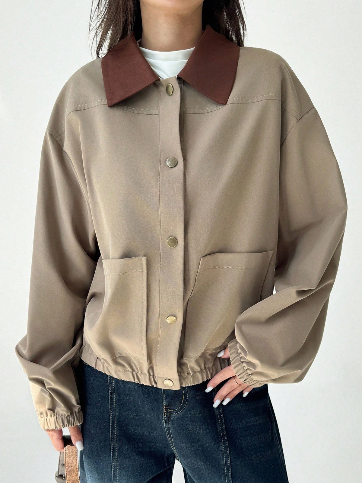 A woman wearing a khaki casual jacket with a contrasting brown lapel collar, drop shoulders, and front pockets paired with a white tee and dark jeans