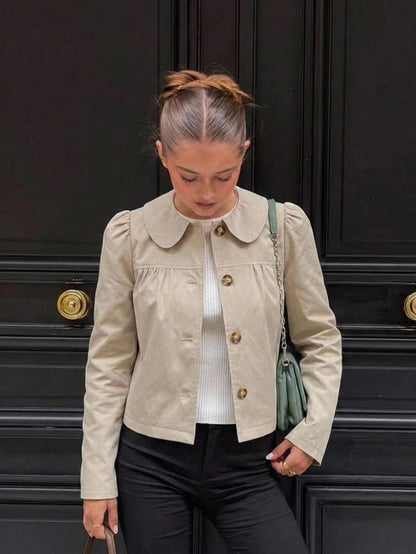A woman wearing a khaki short trench jacket with a Peter Pan collar and tortoiseshell buttons paired with a white top and black pants