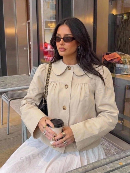 A woman wearing a khaki short trench jacket with a Peter Pan collar and tortoiseshell buttons paired with a white top and black pants