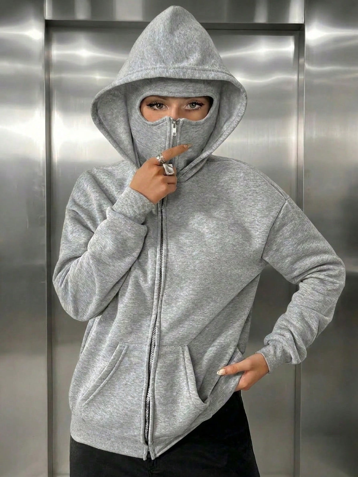 A woman wearing a light grey fleece balaclava zip-up hoodie featuring an integrated face mask and front pockets while standing indoors