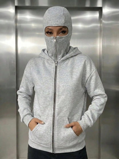 A woman wearing a light grey fleece balaclava zip-up hoodie featuring an integrated face mask and front pockets while standing indoors