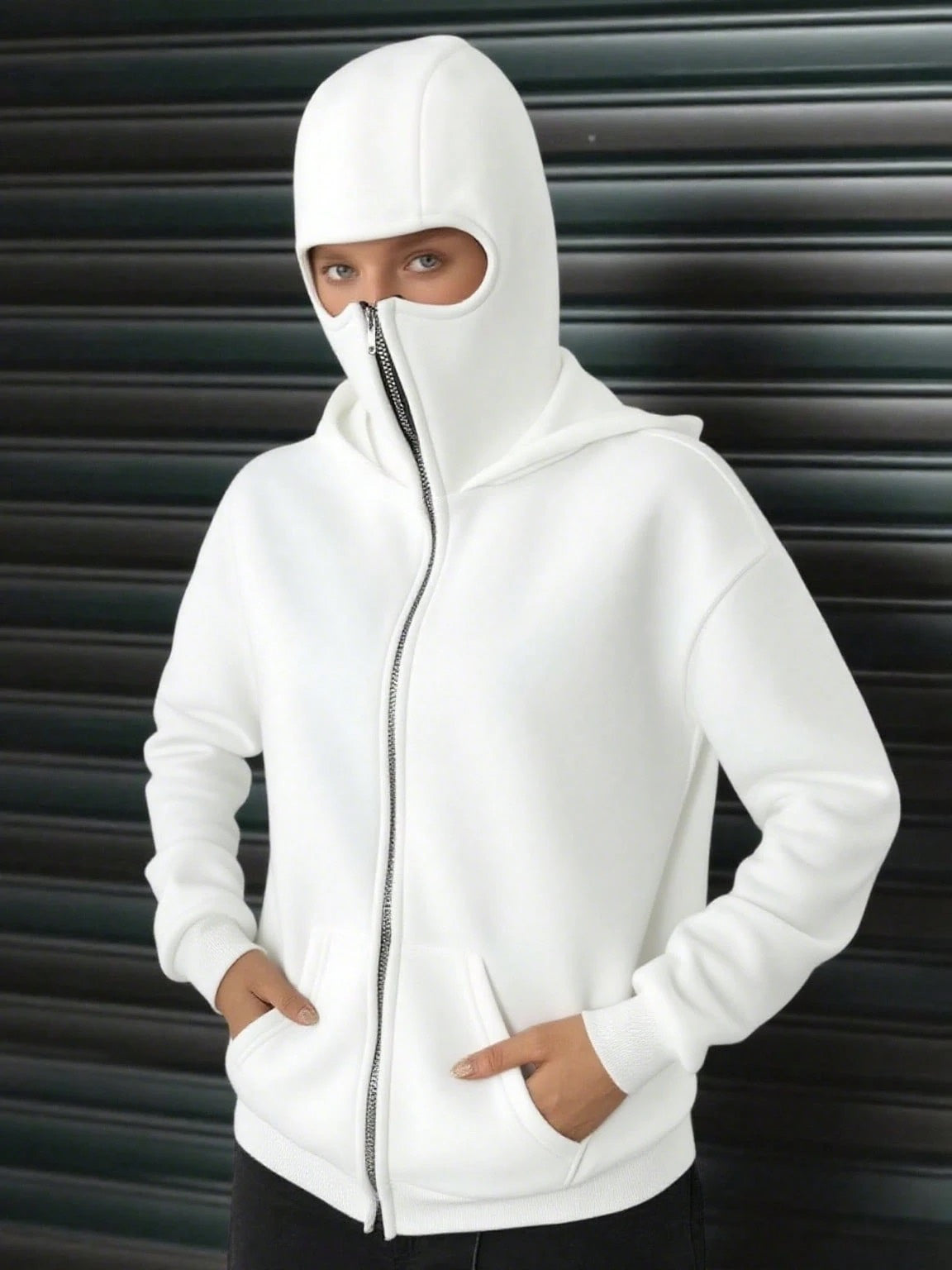 A woman wearing a white fleece balaclava zip-up hoodie featuring an integrated face mask and front pockets while standing indoors
