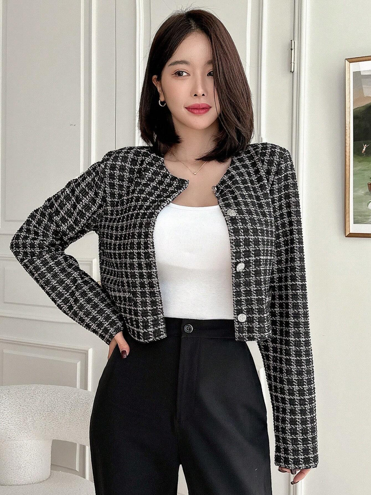 A woman wearing an Black plaid tweed crop jacket featuring a round neckline and long sleeves paired with a pleated skirt