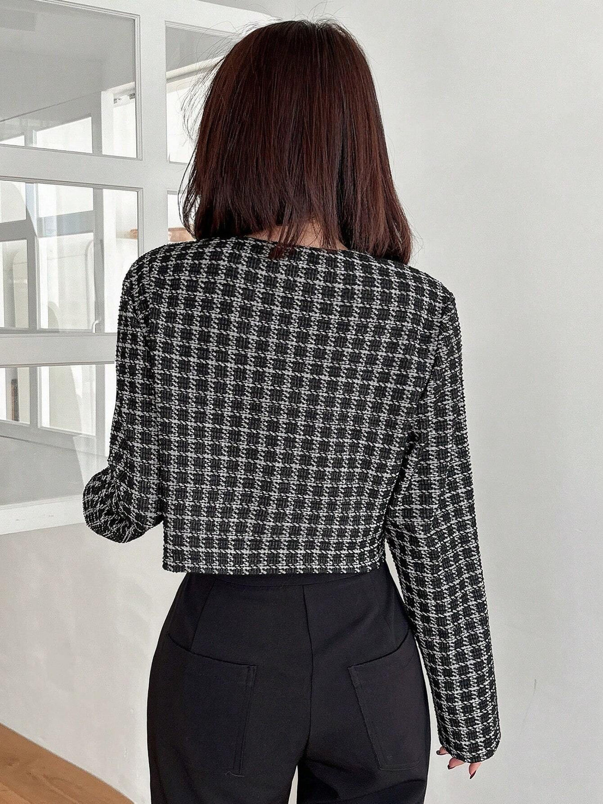 A woman wearing an Black plaid tweed crop jacket featuring a round neckline and long sleeves paired with a pleated skirt