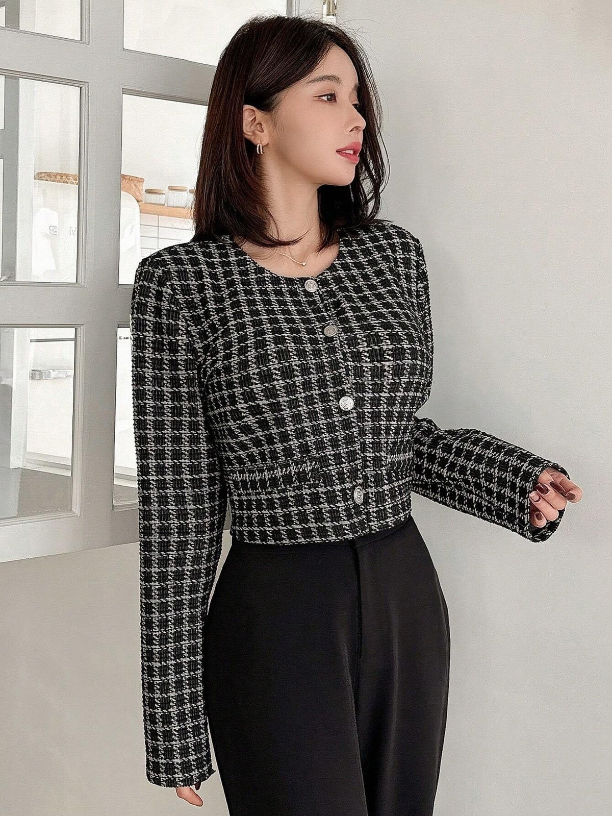 A woman wearing an Black plaid tweed crop jacket featuring a round neckline and long sleeves paired with a pleated skirt