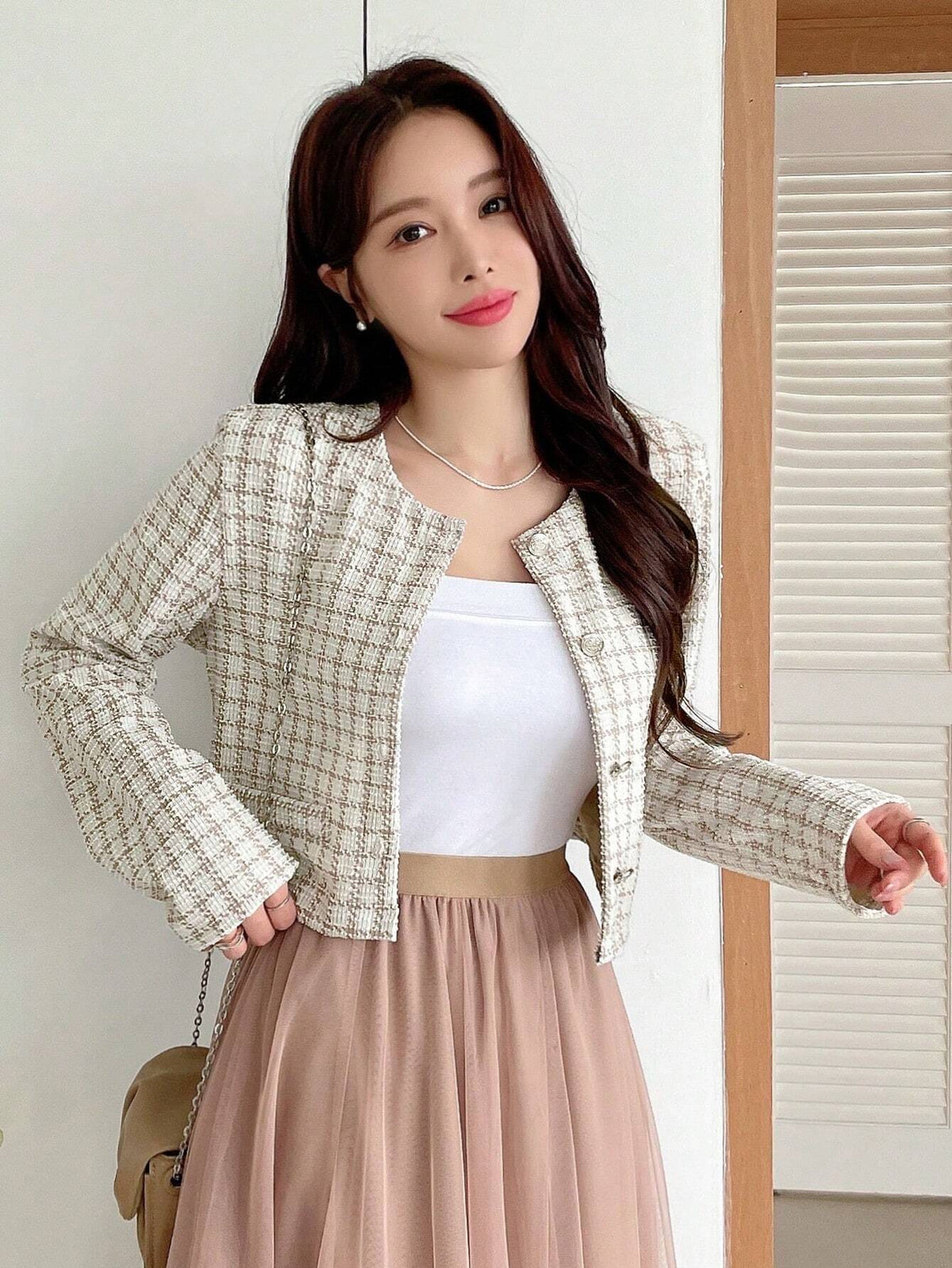 A woman wearing an apricot plaid tweed crop jacket featuring a round neckline and long sleeves paired with a pleated skirt
