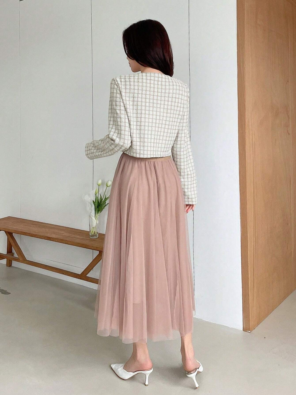 A woman wearing an apricot plaid tweed crop jacket featuring a round neckline and long sleeves paired with a pleated skirt