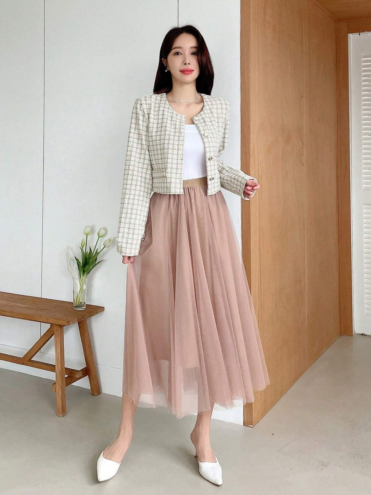 A woman wearing an apricot plaid tweed crop jacket featuring a round neckline and long sleeves paired with a pleated skirt