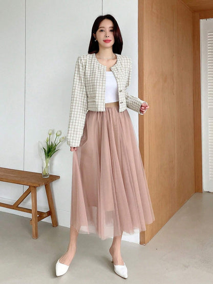 A woman wearing an apricot plaid tweed crop jacket featuring a round neckline and long sleeves paired with a pleated skirt