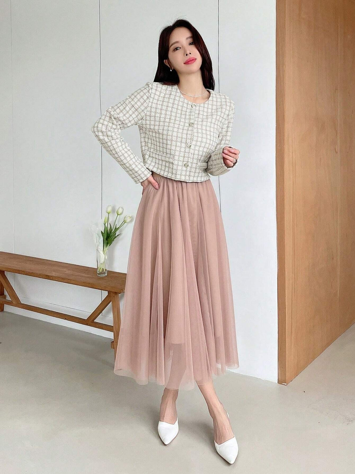 A woman wearing an apricot plaid tweed crop jacket featuring a round neckline and long sleeves paired with a pleated skirt