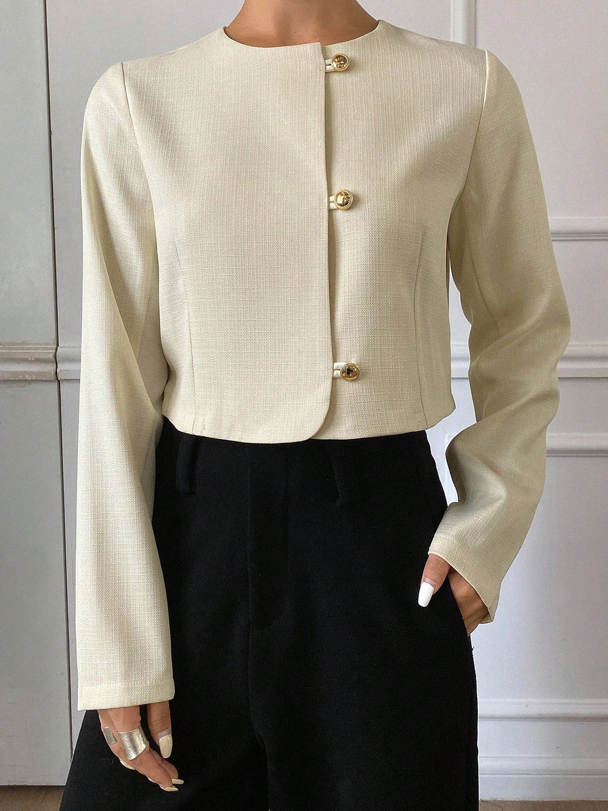 A woman wearing an apricot short jacket featuring gold button accents and long sleeves paired with high-waisted black trousers