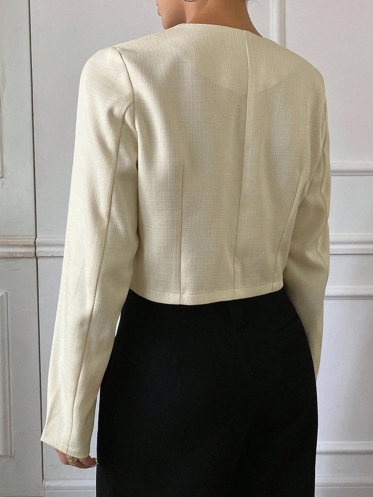 A woman wearing an apricot short jacket featuring gold button accents and long sleeves paired with high-waisted black trousers