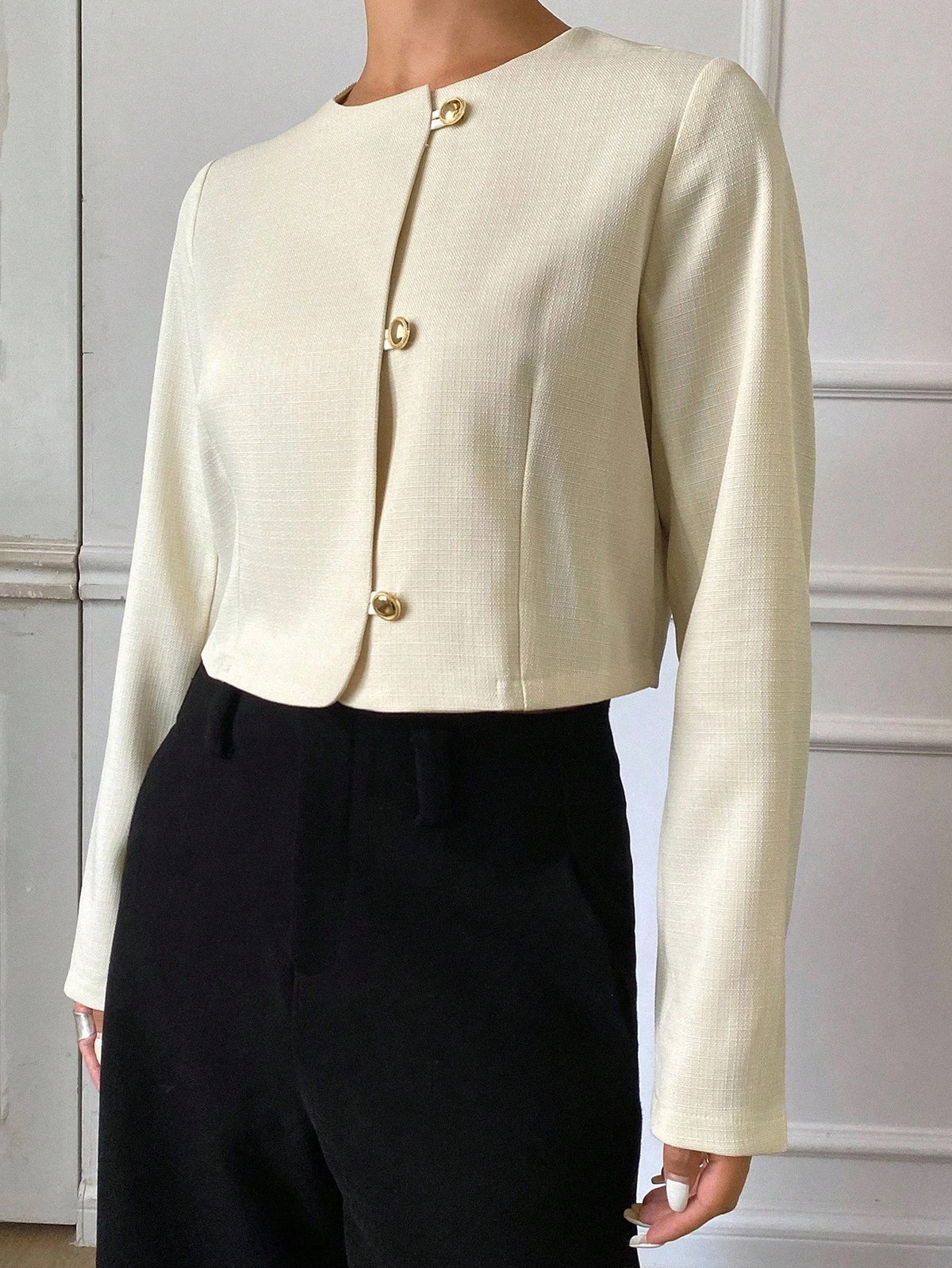 A woman wearing an apricot short jacket featuring gold button accents and long sleeves paired with high-waisted black trousers