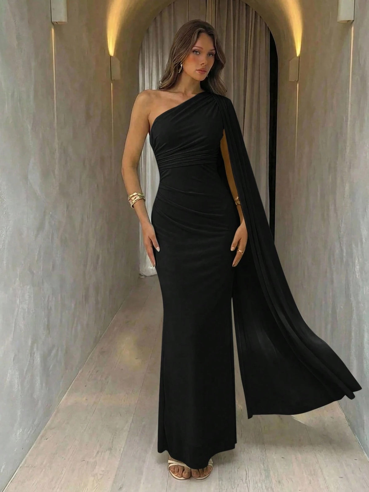 A woman wearing an elegant Black  one-shoulder ruched mermaid long dress with a dramatic flutter cloak sleeve