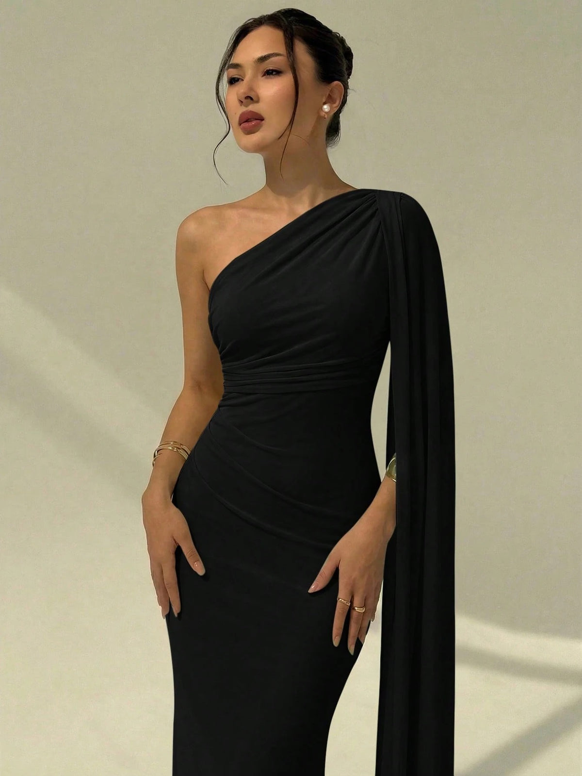 A woman wearing an elegant Black  one-shoulder ruched mermaid long dress with a dramatic flutter cloak sleeve