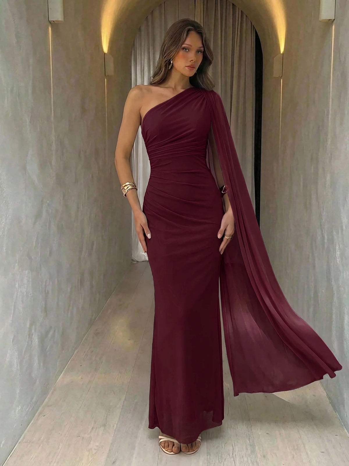 A woman wearing an elegant Burgundy one-shoulder ruched mermaid long dress with a dramatic cloak sleeve in front of a mirror
