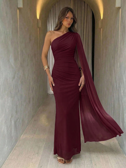 A woman wearing an elegant Burgundy one-shoulder ruched mermaid long dress with a dramatic cloak sleeve in front of a mirror