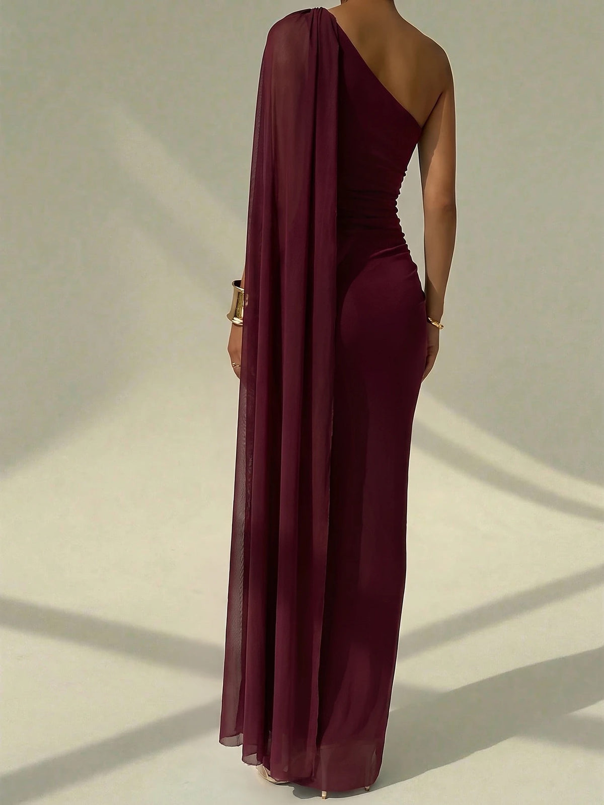 A woman wearing an elegant Burgundy one-shoulder ruched mermaid long dress with a dramatic cloak sleeve in front of a mirror