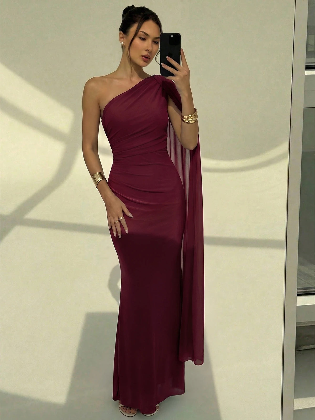 A woman wearing an elegant Burgundy one-shoulder ruched mermaid long dress with a dramatic cloak sleeve in front of a mirror