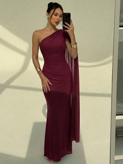 A woman wearing an elegant Burgundy one-shoulder ruched mermaid long dress with a dramatic cloak sleeve in front of a mirror
