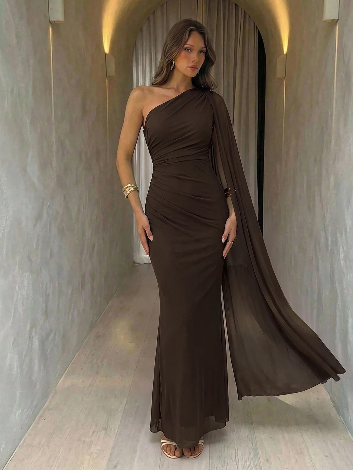 A woman wearing an elegant coffee brown one-shoulder ruched mermaid long dress with a dramatic flutter cloak sleeve