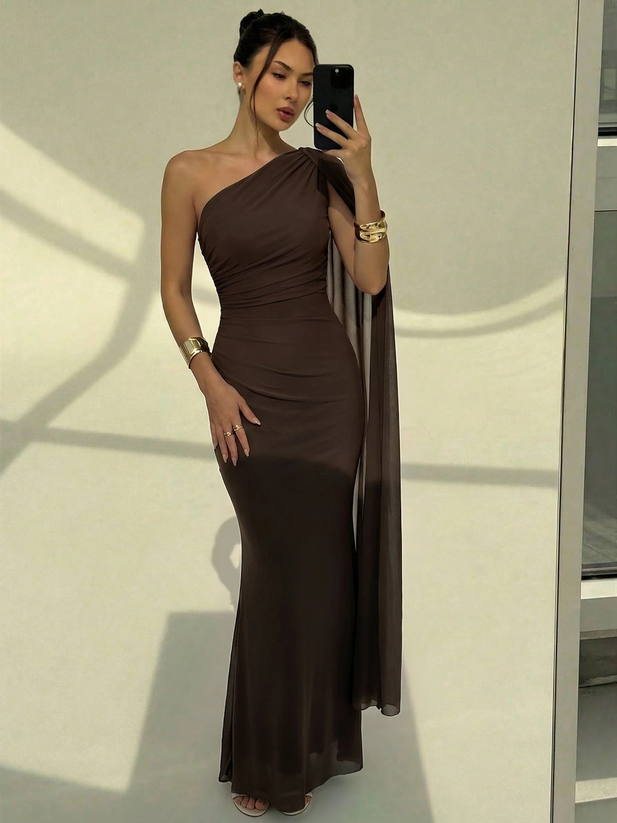 A woman wearing an elegant coffee brown one-shoulder ruched mermaid long dress with a dramatic flutter cloak sleeve