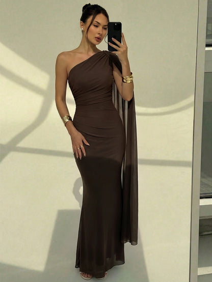 A woman wearing an elegant coffee brown one-shoulder ruched mermaid long dress with a dramatic flutter cloak sleeve
