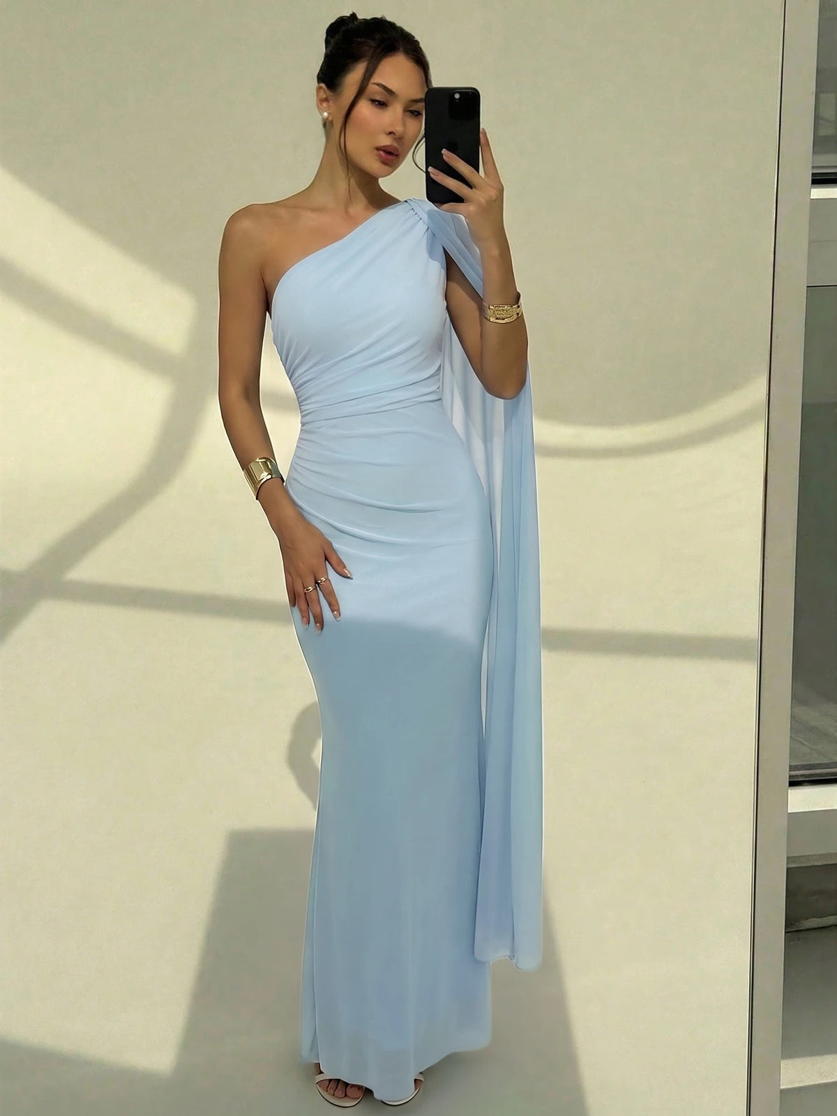 A woman wearing an elegant light blue one-shoulder ruched mermaid long dress with a dramatic flutter cloak sleeve