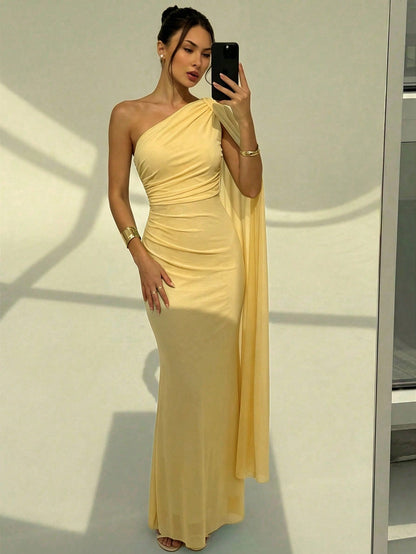 A woman wearing an elegant yellow one-shoulder ruched mermaid long dress with a dramatic cloak sleeve in front of a mirror