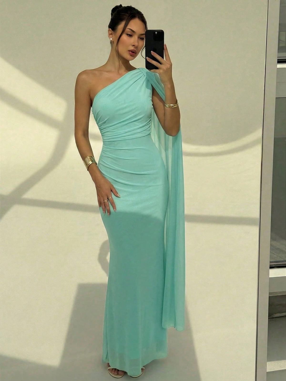 A woman wearing an elegant Mint Dress one-shoulder ruched mermaid long dress with a dramatic flutter cloak sleeve