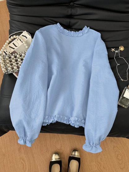 Light blue frill trim tie-front long-sleeve blouse with delicate ruffles laid flat on a dark textured background