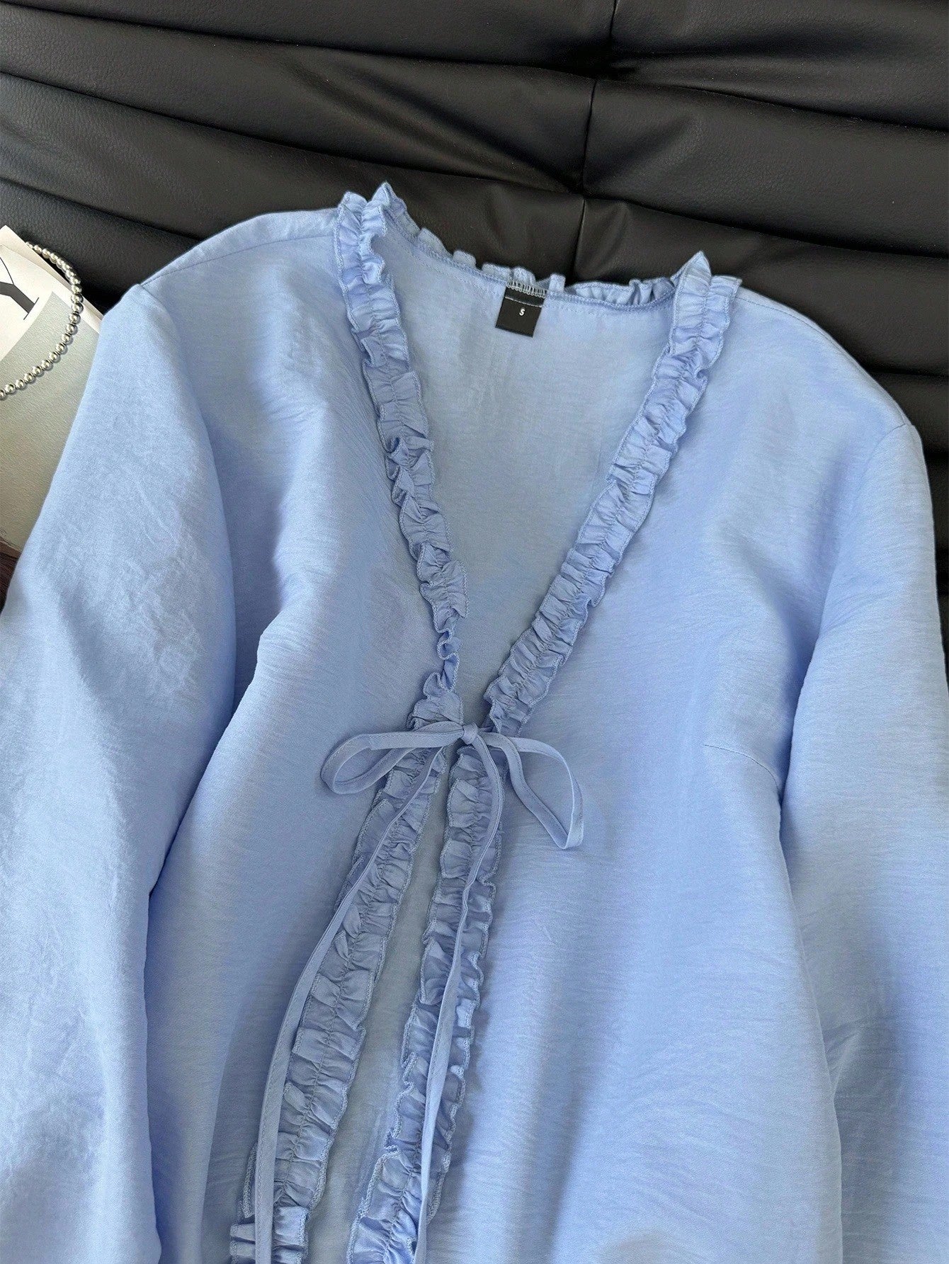 Light blue frill trim tie-front long-sleeve blouse with delicate ruffles laid flat on a dark textured background