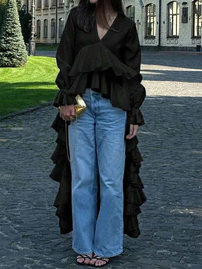 Woman wearing a black long-sleeve V-neck top with an asymmetrical cascading ruffle high-low hem paired with blue jeans and holding a gold clutch.