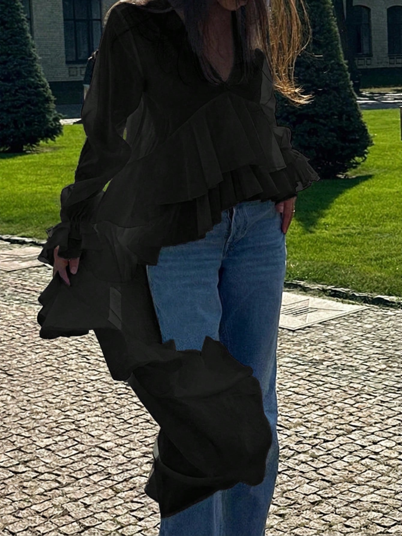 Woman wearing a black long-sleeve V-neck top with an asymmetrical cascading ruffle high-low hem paired with blue jeans and holding a gold clutch.