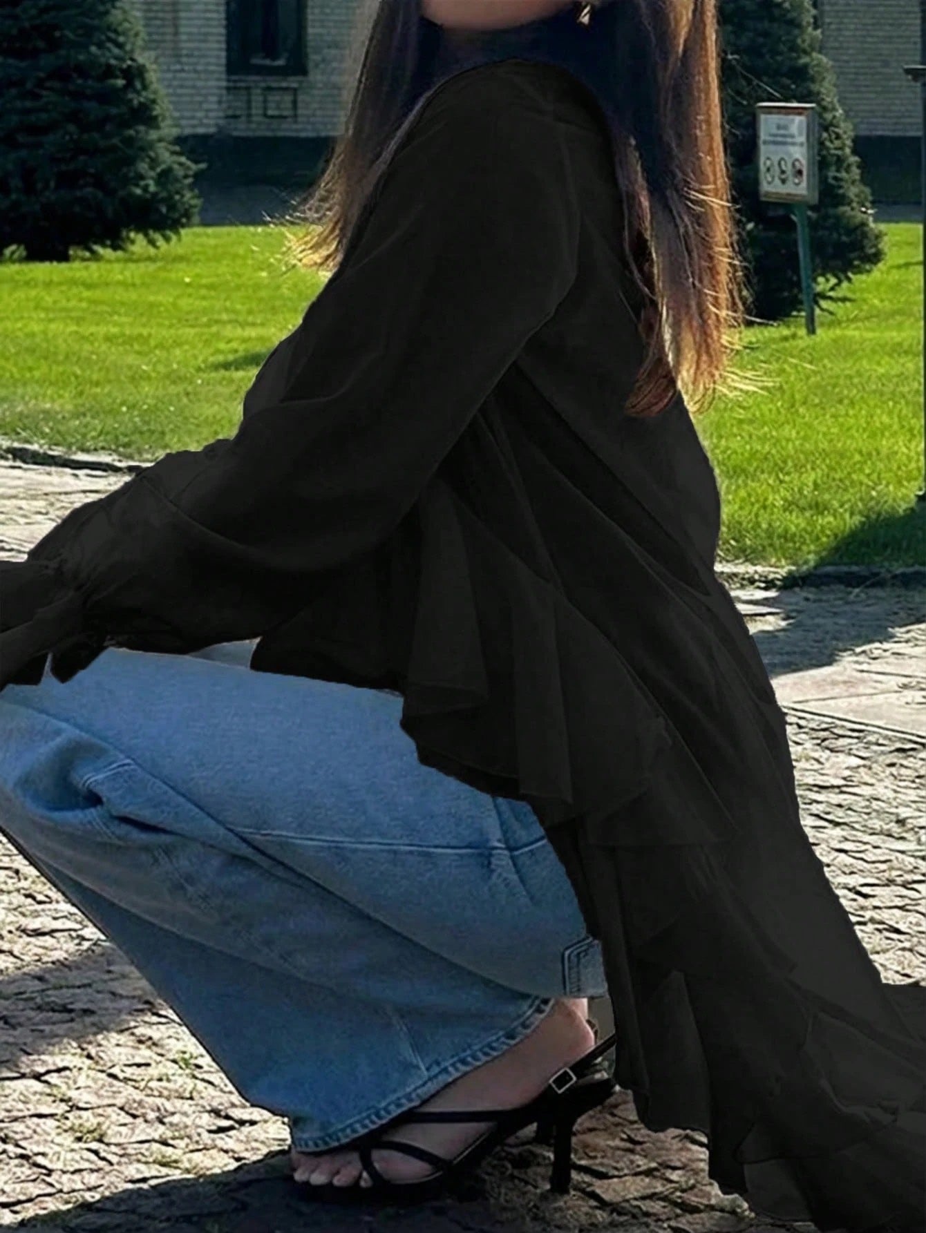 Woman wearing a black long-sleeve V-neck top with an asymmetrical cascading ruffle high-low hem paired with blue jeans and holding a gold clutch.
