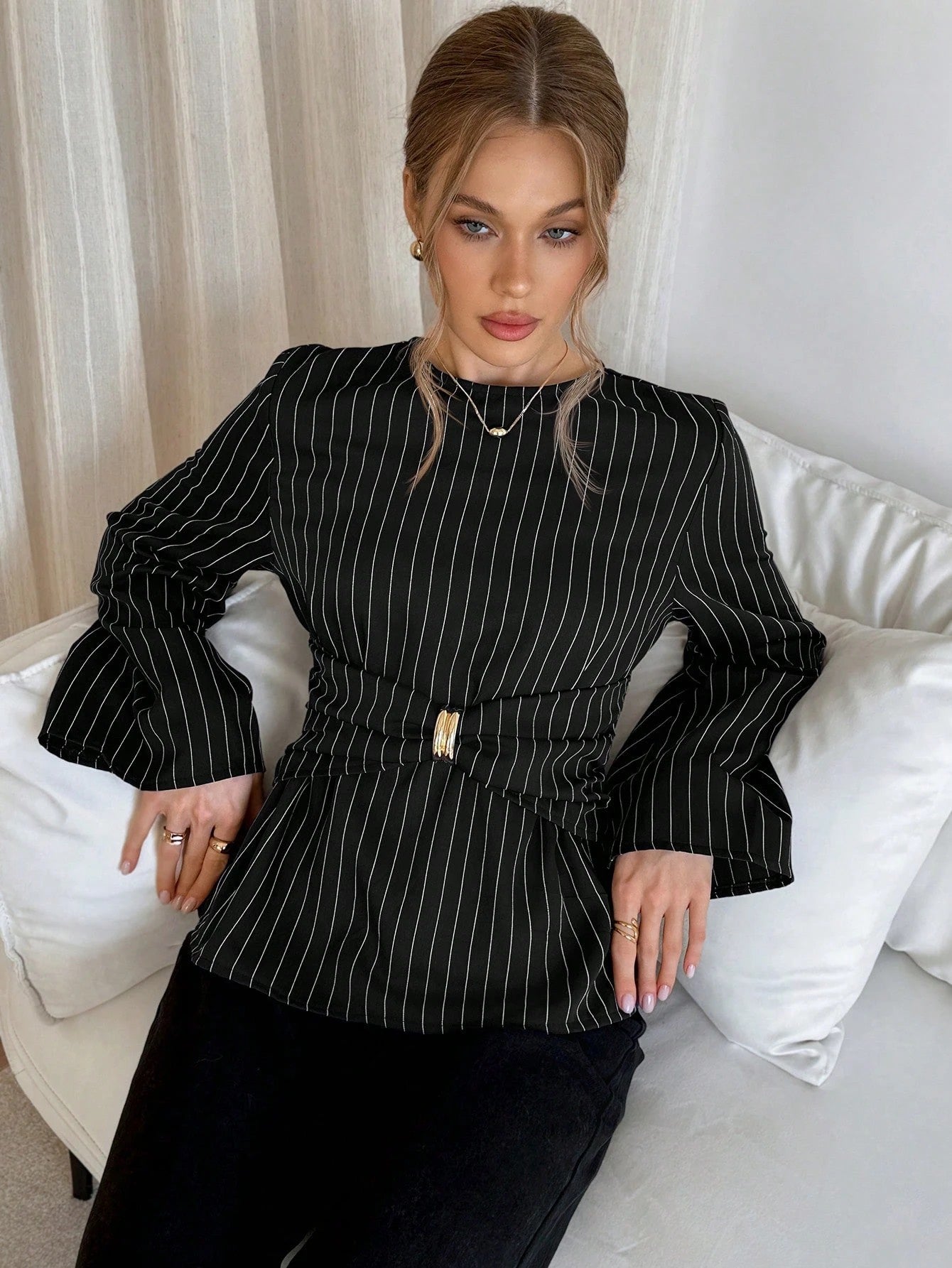 Woman wearing a black pinstripe long sleeve blouse featuring a metal ring decor waist detail and dark trousers