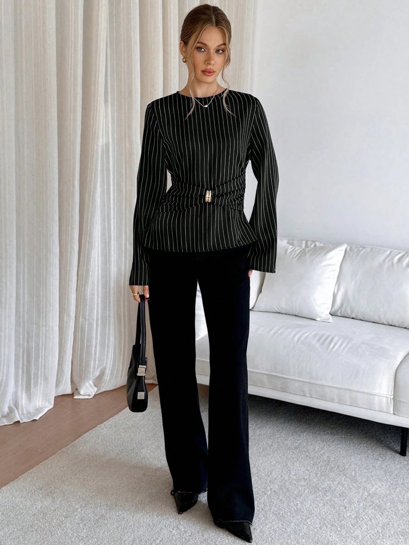 Woman wearing a black pinstripe long sleeve blouse featuring a metal ring decor waist detail and dark trousers