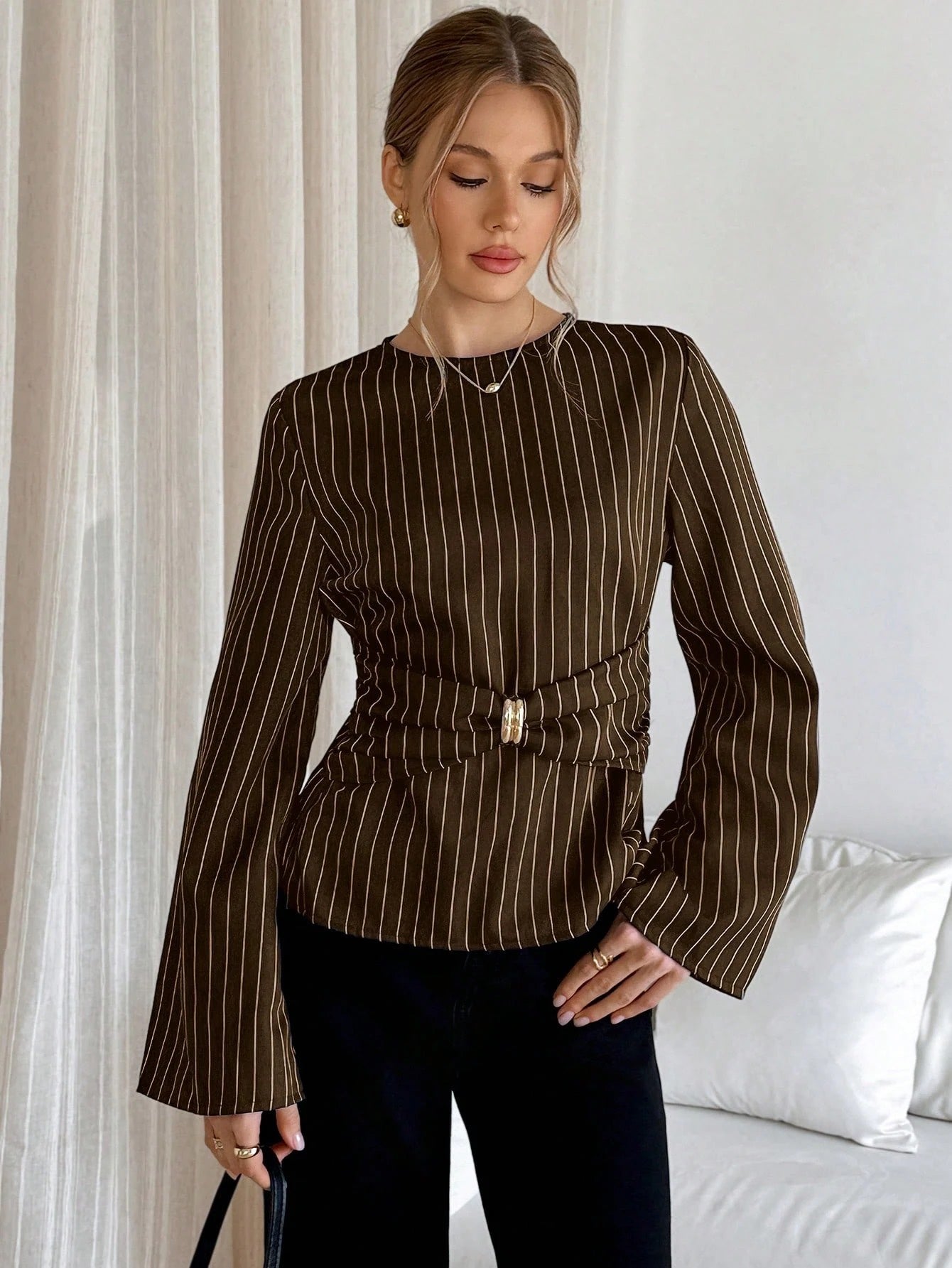 Woman wearing a brown pinstripe long sleeve blouse featuring a metal ring decor waist detail and dark trousers
