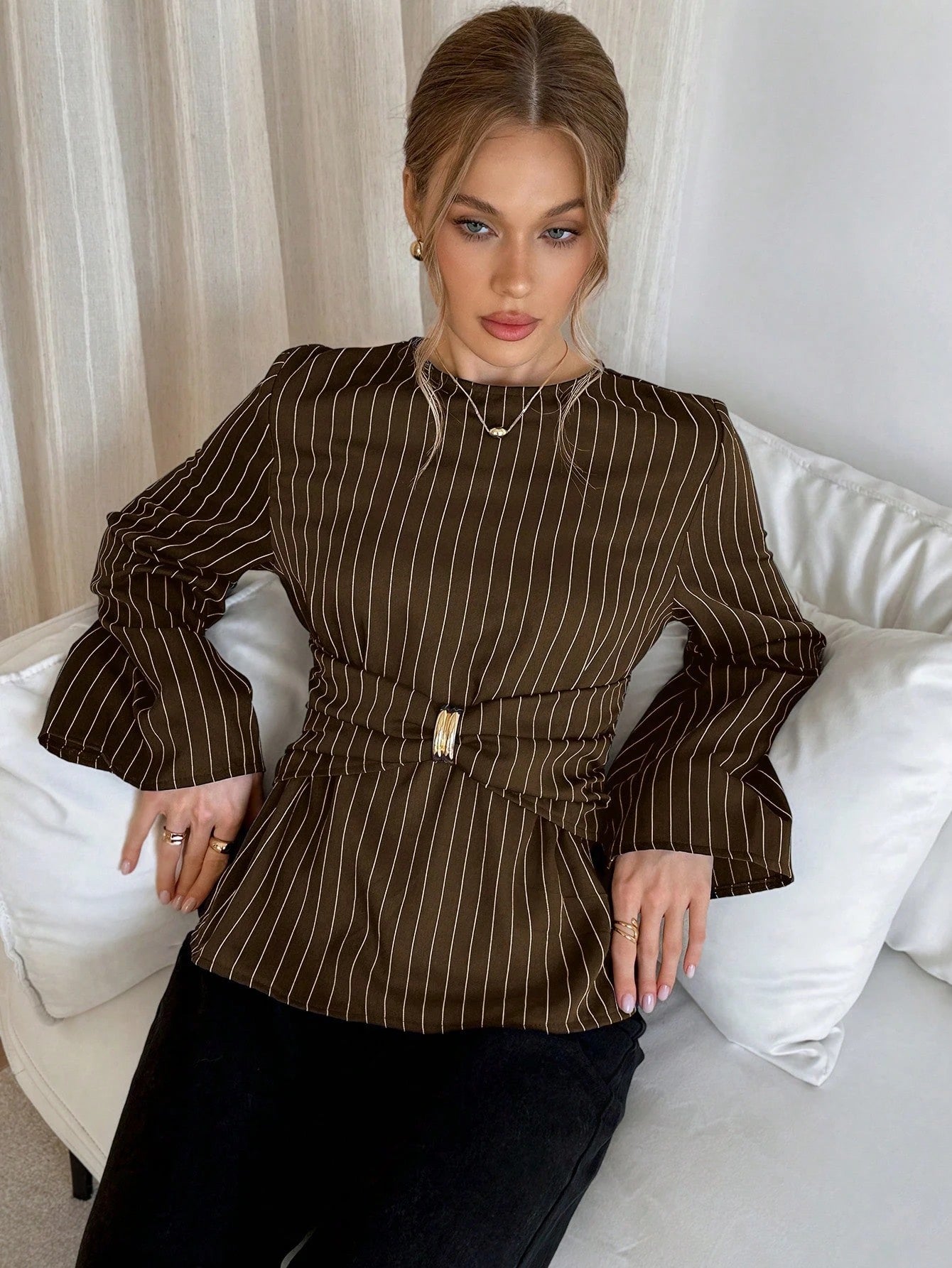 Woman wearing a brown pinstripe long sleeve blouse featuring a metal ring decor waist detail and dark trousers