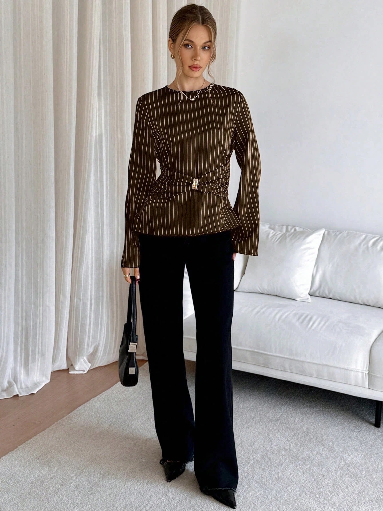 Woman wearing a brown pinstripe long sleeve blouse featuring a metal ring decor waist detail and dark trousers