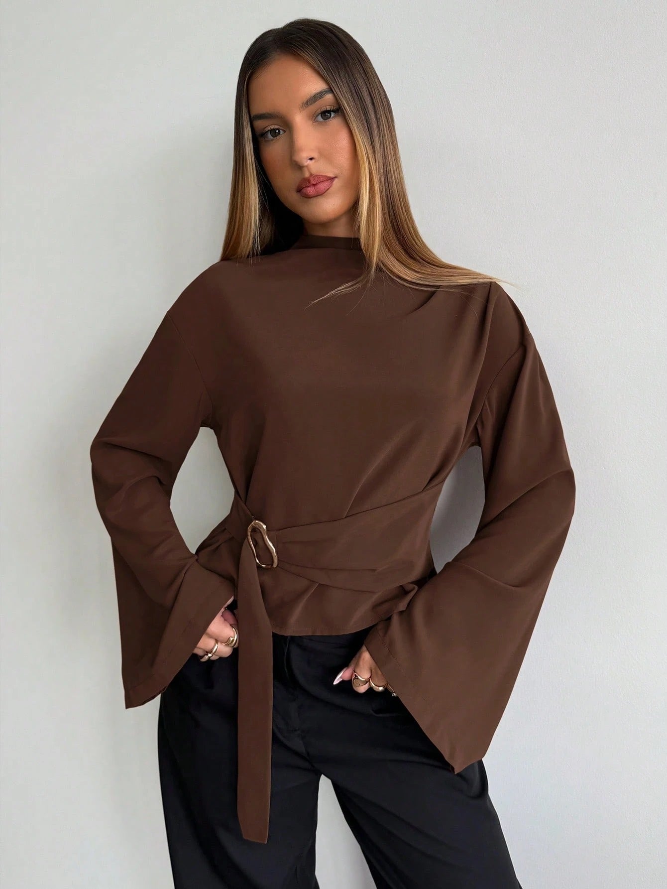 Woman wearing a coffee brown mock-neck long-sleeve top with bell sleeves and an integrated waist belt with a gold buckle against a white wall.